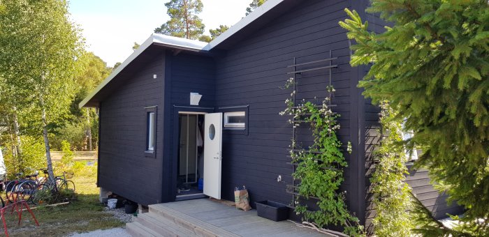 Svart trähus med vitt tak och vita fönsterkarmar, trätrall framför huset, klätterväxt på fasaden.