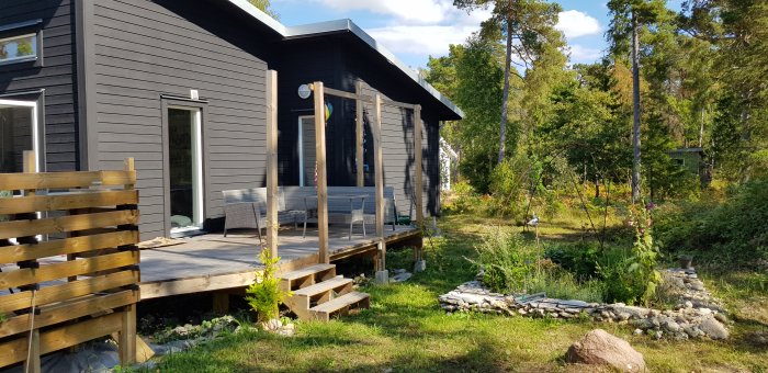 Ett färdigt byggprojekt av ett modernt grått hus med terrass och naturskön omgivning i solljus.