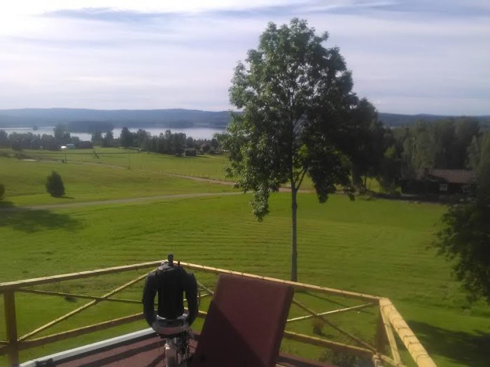 Utsikt från ett observationsdäck med en person, gröna fält, träd och en sjö i bakgrunden.