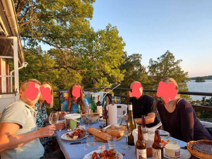 Personer som njuter av en middag utomhus med kräftor och dryck på en terrass med utsikt över vatten och grönska i solnedgången.