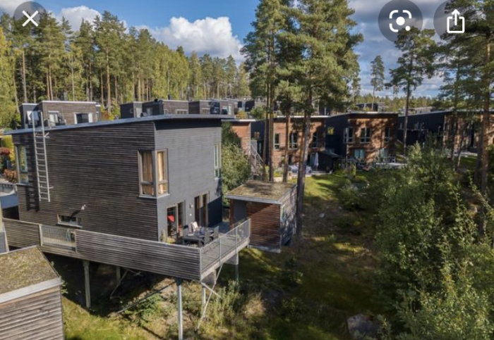 Modern flervåningshus med träfasad, balkonger och en upphöjd terrass, omgivna av skog.