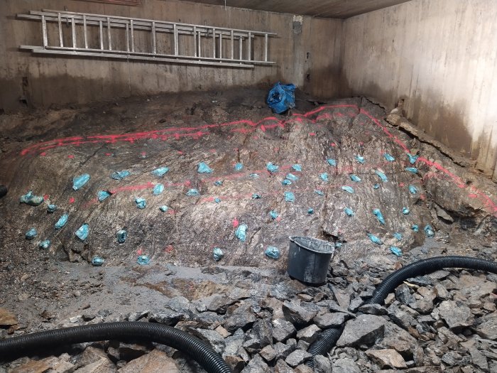 Grund med borrade hål markerade med blå plast och röd sprayfärg, stenhög och dräneringsslangar.