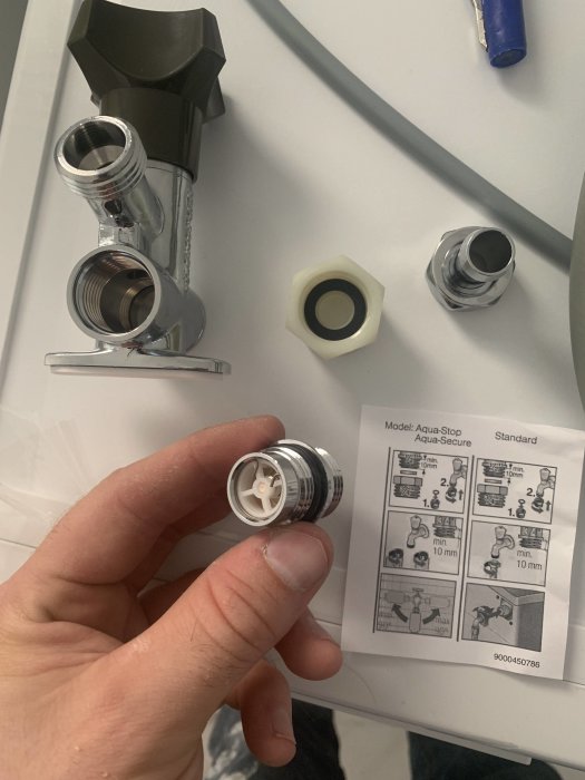 Hand håller en Aqua Stop Secure tappventil över instruktionsblad med rörmokarverktyg i bakgrunden.