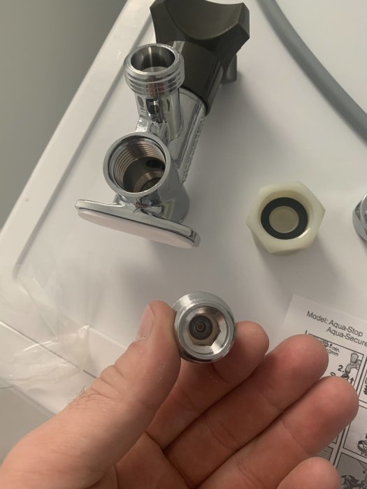Hand håller en liten metallkomponent framför en kran och andra VVS-tillbehör.