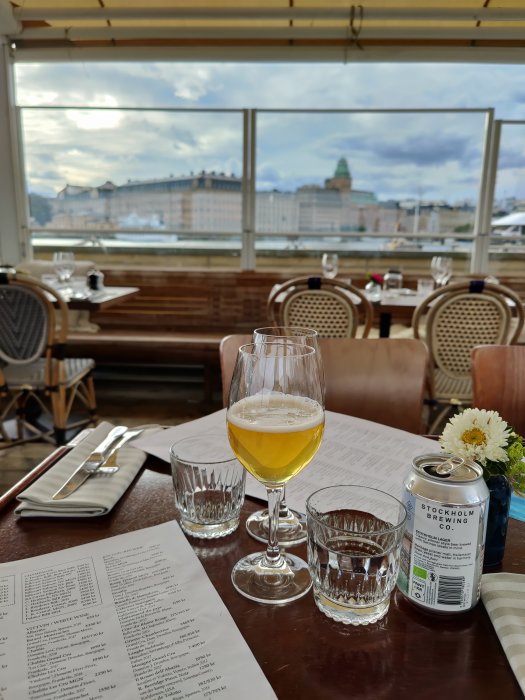 Ölglas och burk på bord med meny framför fönster med utsikt över stad och molnig himmel.