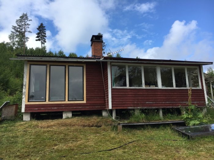 Rödmålad stuga med lutning och murstock, stöttad av blandade plintar av lecablock och tegelstenar.