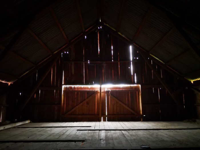 Mörkt stallloft med ljusinflöde genom sprickor i dörrar och tak, föreslagen för ombyggnad till lägenhet.