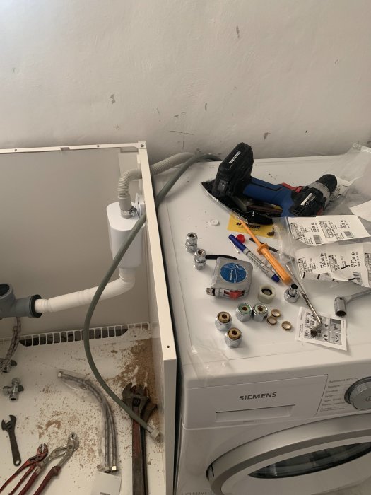 Tvättmaskinsinstallation med avloppsslang och verktyg synliga på ett stökigt golvet.