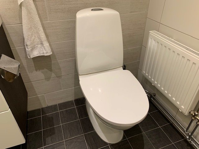 Vit toalettstol i hörn av badrum med kaklade väggar, nära handduk och radiator.