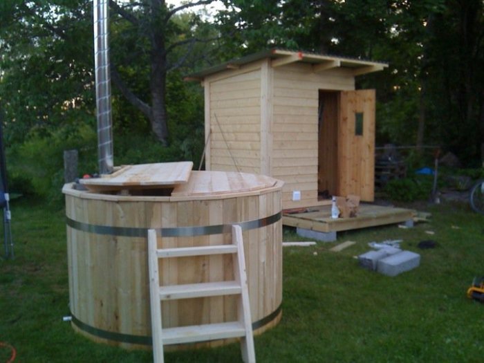 Nybyggd bastu av trä med skorsten och intilliggande omklädningsrum vid skogsbryn.