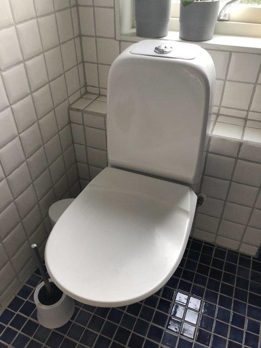 Vägghängd vit toalett från Gustavsberg med stängd sits i ett badrum med kakel och klinkergolv.