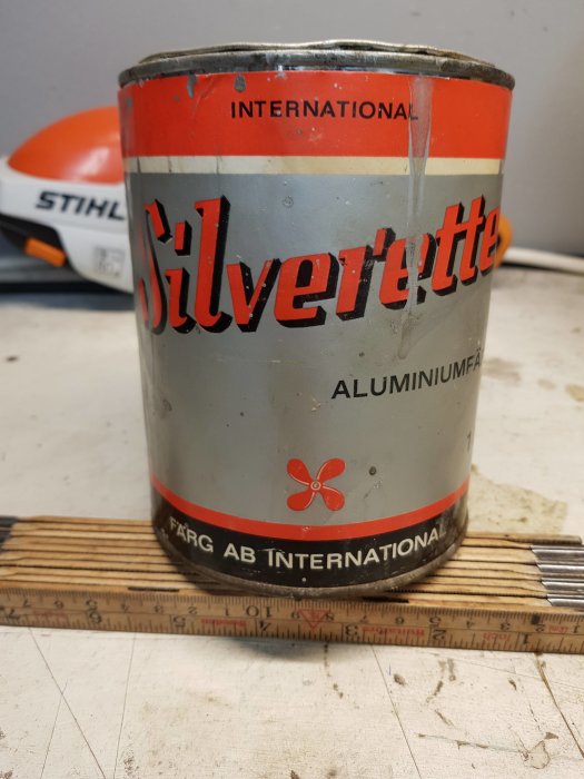 En burk Silverette aluminiumfärg på ett arbetsbord med måttstock, bakom syns del av en STIHL hjälm.