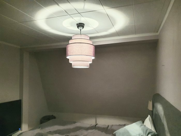 Interiörbild på sovrum med synlig pelare och del av balk i taket, samt en taklampa och säng.