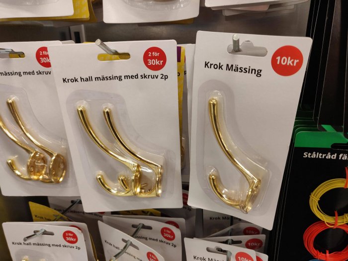 Förpackningar med guldfärgade krokar märkta "2 för 30kr" och "Krok Mässing 10kr" i en butikshylla.