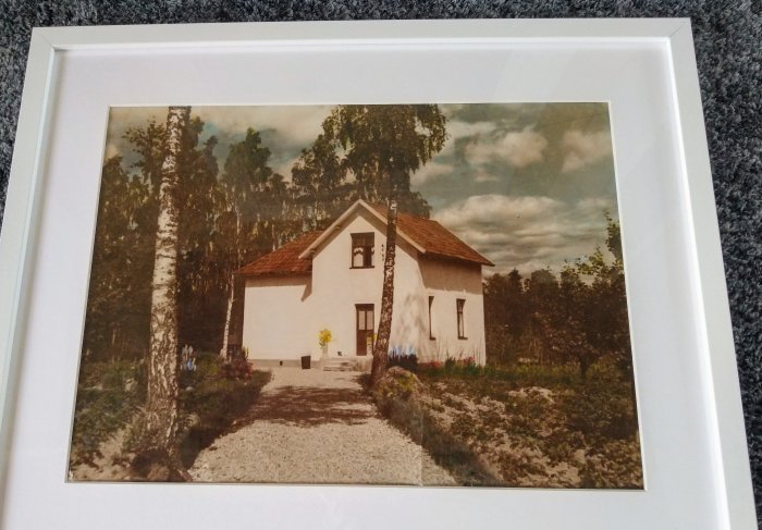 Inramat foto av vitt hus med två våningar från 1940-talet, omgivet av natur.