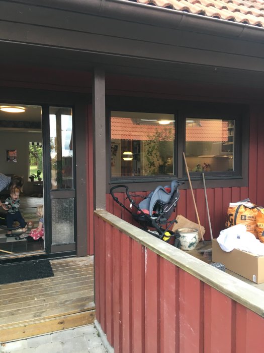 Ulko-oven edessä on puinen pylväs yhdistettynä kattoon. Lapsia leikkimässä sisällä, lastenrattaat, rakennusmateriaalia ja istutusastia verannalla.