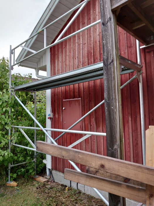 Nybyggd ställning av metall vid en rödmålad garagevägg med öppen dörr och molnig himmel.