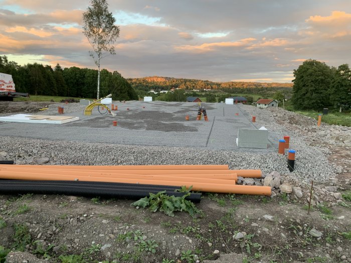 Byggplats med grus, avgjord område med markeringskoner och orangea avloppsrör i förgrunden.