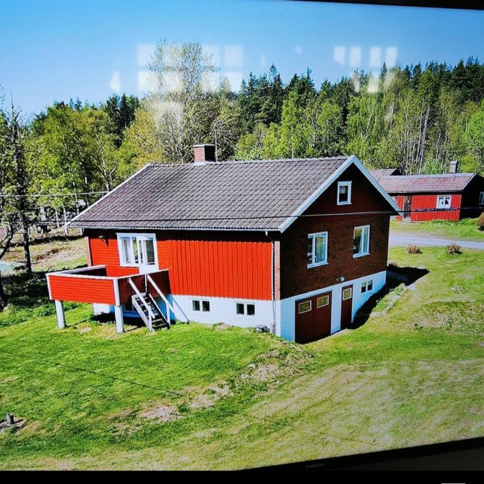 Tvåvåningshus med röd fasad på nedervåningen och tegelbrun ovanför, omgivet av grönt gräs och träd.