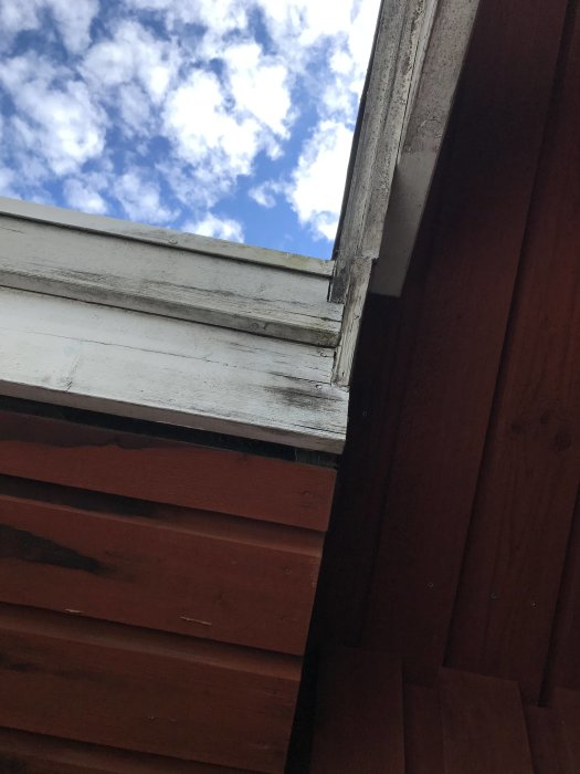 Vy från under en balkong med fuktmarkerad takbjälke och vindskivor mot en bakgrund av blå himmel och moln.