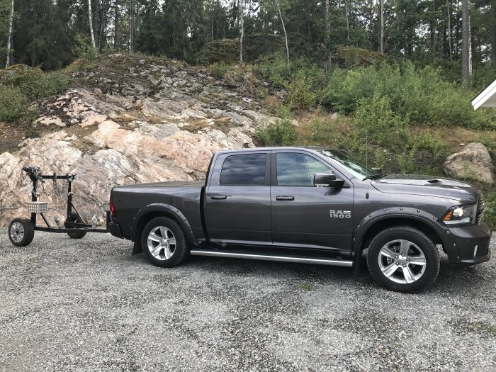 Svart pickup med en lunningsvagn kopplad bak, parkerad framför ett skogsbeklätt berg.