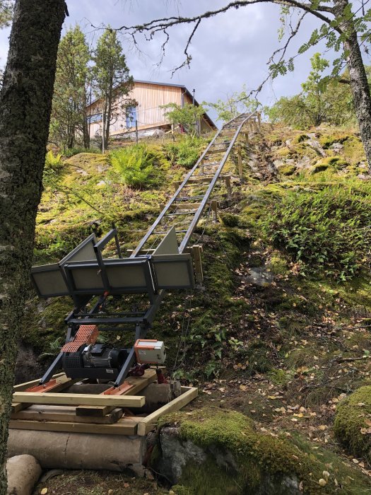 Trädgårdshiss med vajerbrottskydd på en kuperad terräng leder upp till en röd stuga.