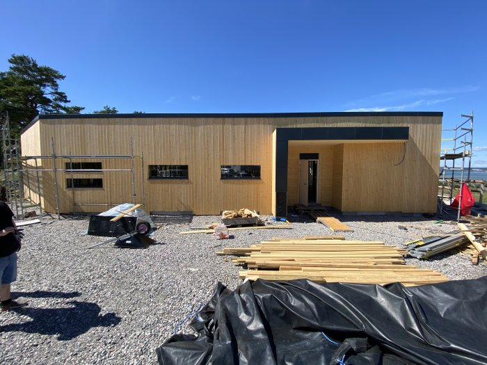 Nybyggd villa med träfasad och byggmaterial framför huset under en klarblå himmel.