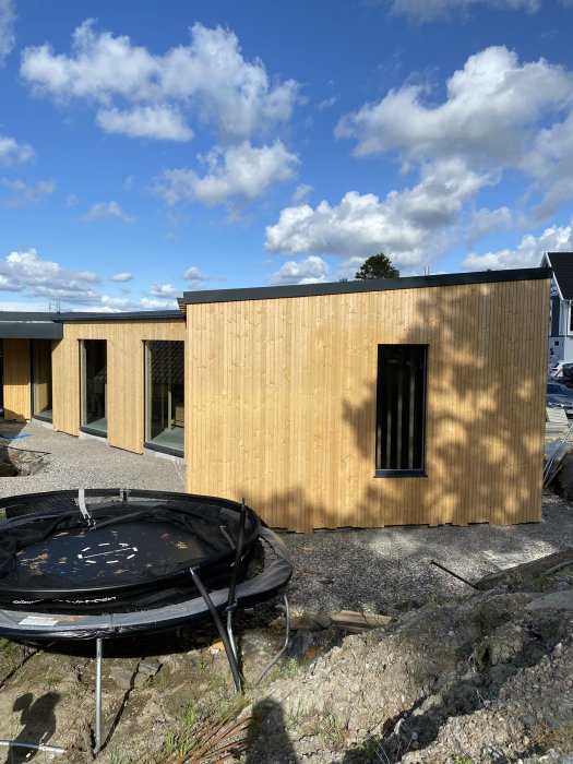 Nybyggt hus med träfasad och stora fönster under blå himmel, omonterad trampolin i förgrunden.