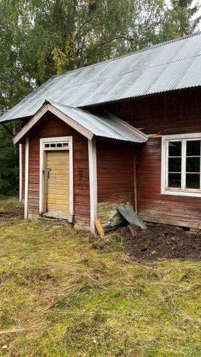 Ett slitet trätorp med skiffertak och en trasig grund i behov av renovering omgivet av träd och gräs.