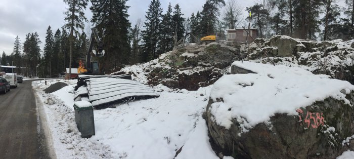 Byggplats med grävmaskin, snötäckta schaktmassor och märkt berg i ett vinterlandskap.