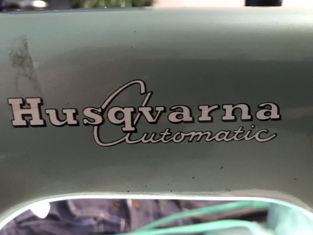 Närbild på logotypen "Husqvarna Automatic" på en grönmetallisk yta, antydan till shorts i bakgrunden.