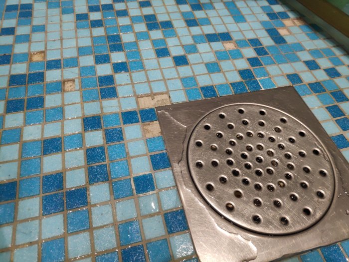 Badrumsgolv med blå mosaikplattor där några plattor saknas nära en silverfärgad golvbrunn.