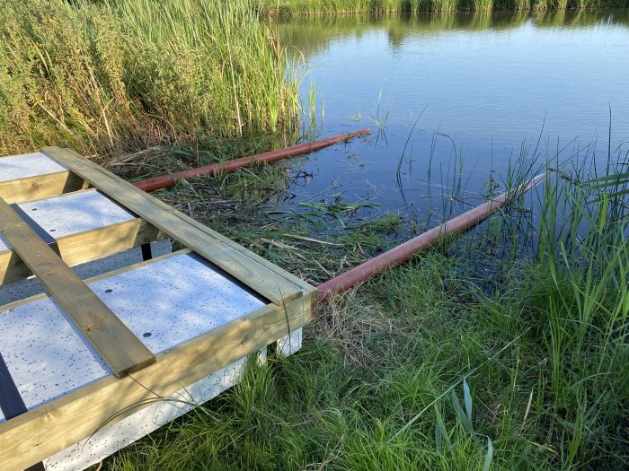 Del av en flytbrygga under konstruktion med cellplastblock och träreglar bredvid en sjö.