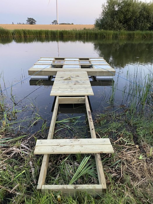 En halvfärdig flytbrygga och gångbro med trall under konstruktion vid en sjö i ett lantligt landskap.