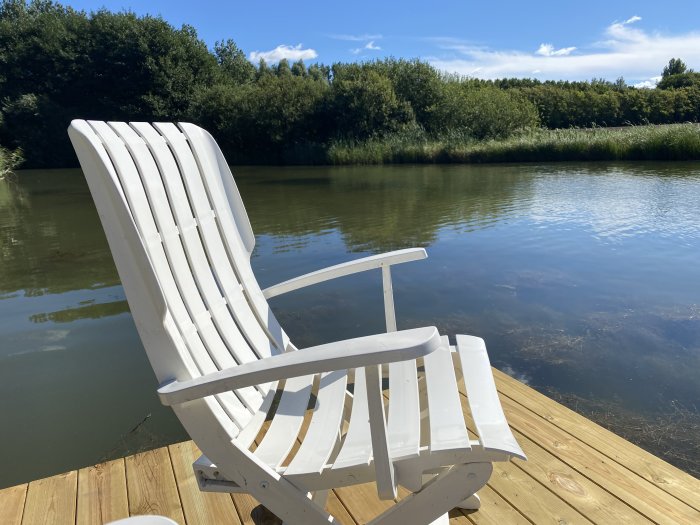 Vit Adirondack-stol på nyskapad trädäckad flytbrygga vid en damm med träd i bakgrunden.