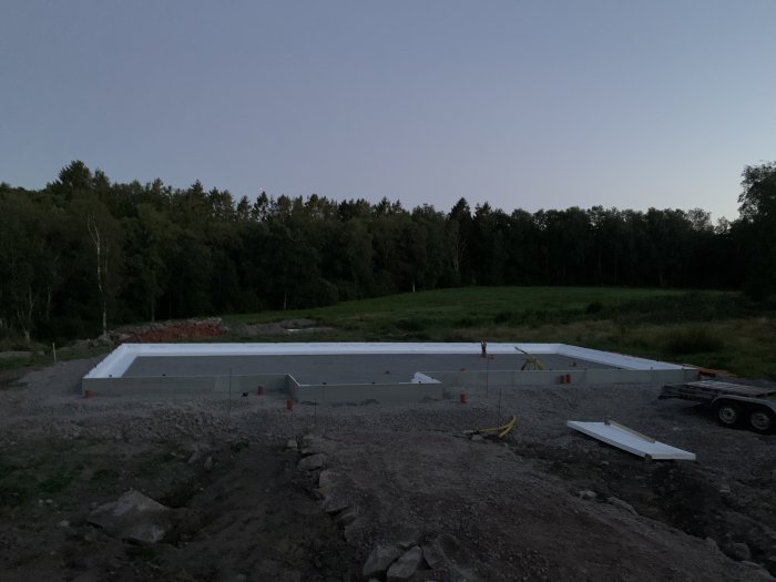 Grundkantelement uppställda på byggarbetsplats i skymning med skog i bakgrunden och släpvagn i förgrunden.