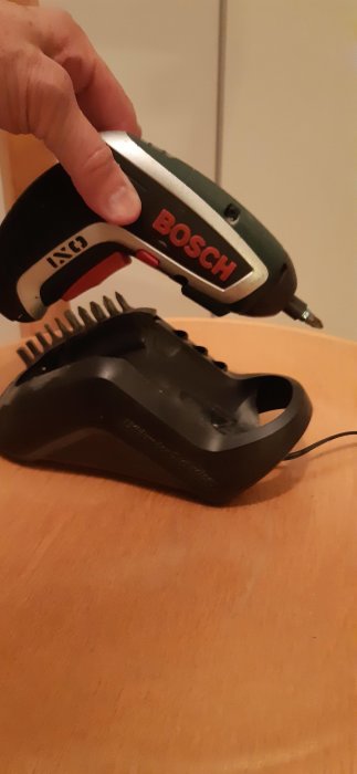 En hand håller en Bosch sladdlös skruvdragare ovanför dess laddningsstation på ett träbord.