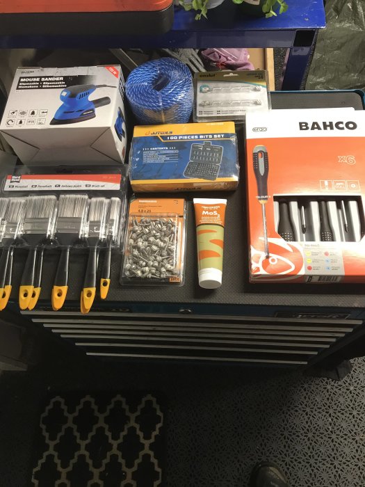 Verktyg och byggmaterial på ett arbetsbord, inklusive en muslipmaskin, rep, skruvmejselsats och en snösläde i förgrunden.