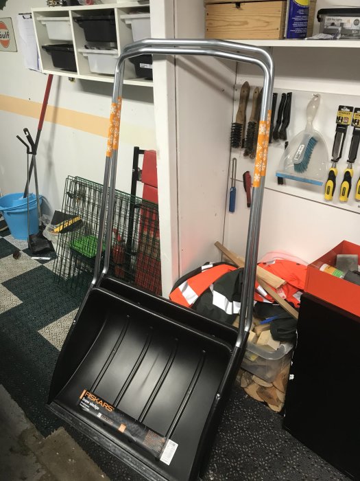 En svart snösläde av märket Fiskars står i ett organiserat garage med verktyg och förvaringshyllor i bakgrunden.