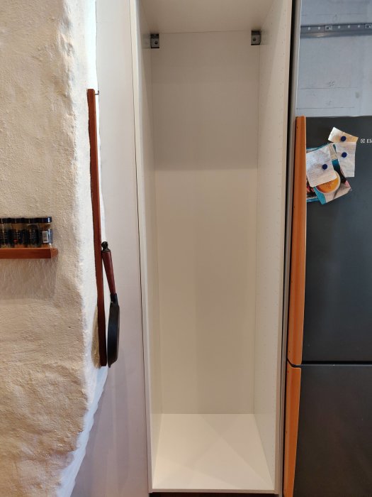 Ett upphängt tomt skåp från IKEA, precis vid en vitmålad vägg, med synliga upphängningsskenor och närliggande kylskåp.