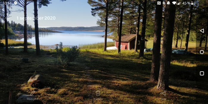 Utsikt från skogskanten över en vacker sjö med klippor, ett rött hus och båtar, omgiven av grönska.