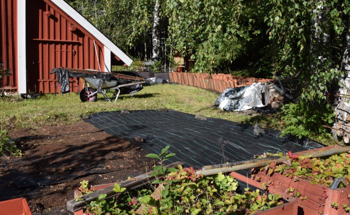 Arbetsområde i trädgård med startat projekt, marktäckning, skottkärra och upplag av tegelpannor.