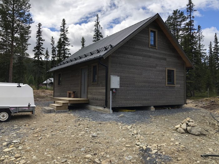 Nybyggd stuga med mörkt träslag och taktäckning, omgiven av skog och grusväg framför.