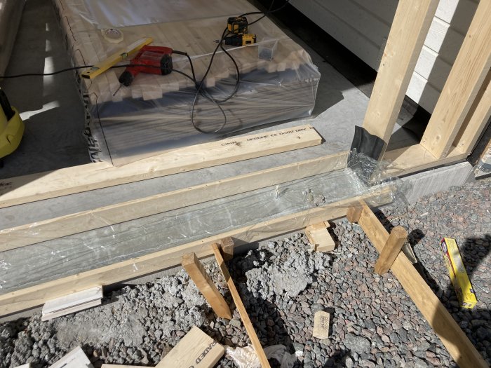 Byggprojekt med nyligen gjuten betongklack, plastfolie i botten, verktyg och böjda bultexpander nära syll.