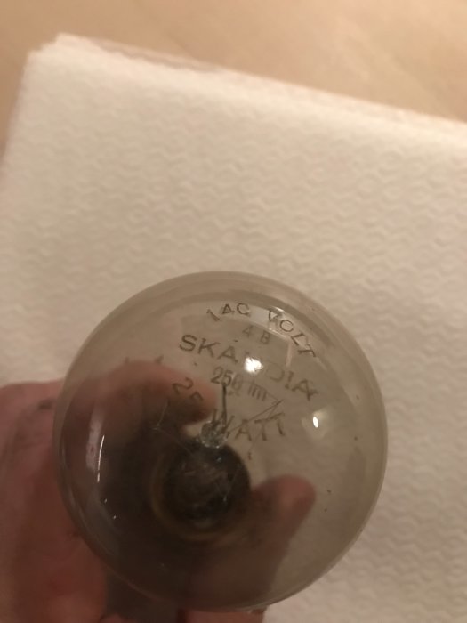 Hand håller en gammal 140V glödlampa med texten "AC VOLTS 110-125 SKANDIA 25 WATT" synlig på glaskulan.