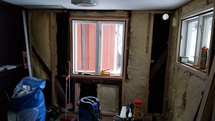 Renoveringsarbete i rum med isolering synlig, två fönster och byggverktyg utspridda.