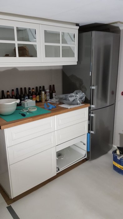 Kök med vit skåpinredning och kylskåp före färdigställande, diverse byggmaterial på bänkskivan.