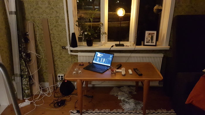 Vardagsrum med retro tapet, bord med laptop och sladdar, fönster med växter och lampa samt soffhörn.