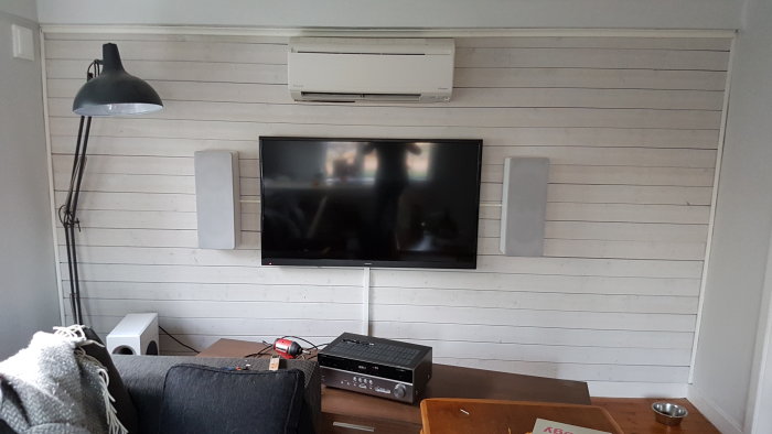 Vardagsrum med väggmonterad TV, högtalare, lampa, och luftkonditionering mot vit träpanelvägg.