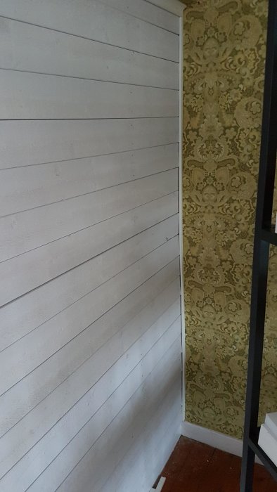 Hörn av vardagsrum med vit träpanelvägg bredvid en vägg med dekorativt mönstrad tapet.